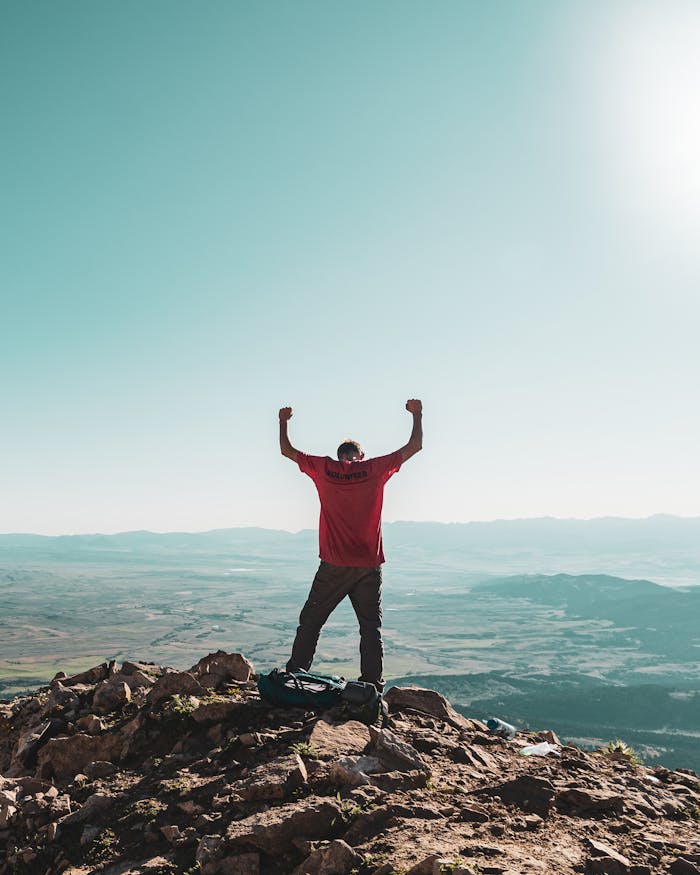 pexels-photo-3680108 A man triumphantly celebrates on a rocky mountain summit, overlooking vast landscapes under clear skies.