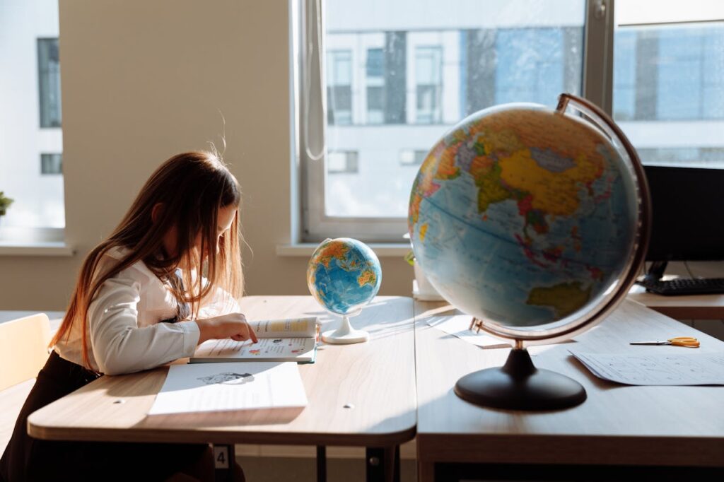 pexels-photo-8926410 A young girl diligently studying in a sunlit classroom with a globe and books.